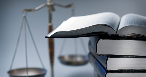 A stack of law books stands in front of a justice scale that is slightly out of focus.  On top of the stack is an open law book.